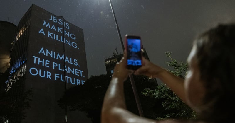 Projections lit up on a high storey building. Text reads: JBS is making a killing. Animals. The Planet. Our Future. A person is taking a photo of it on their mobile phone. Image credit: World Animal Protection Brazil /  Proteção Animal Mundial